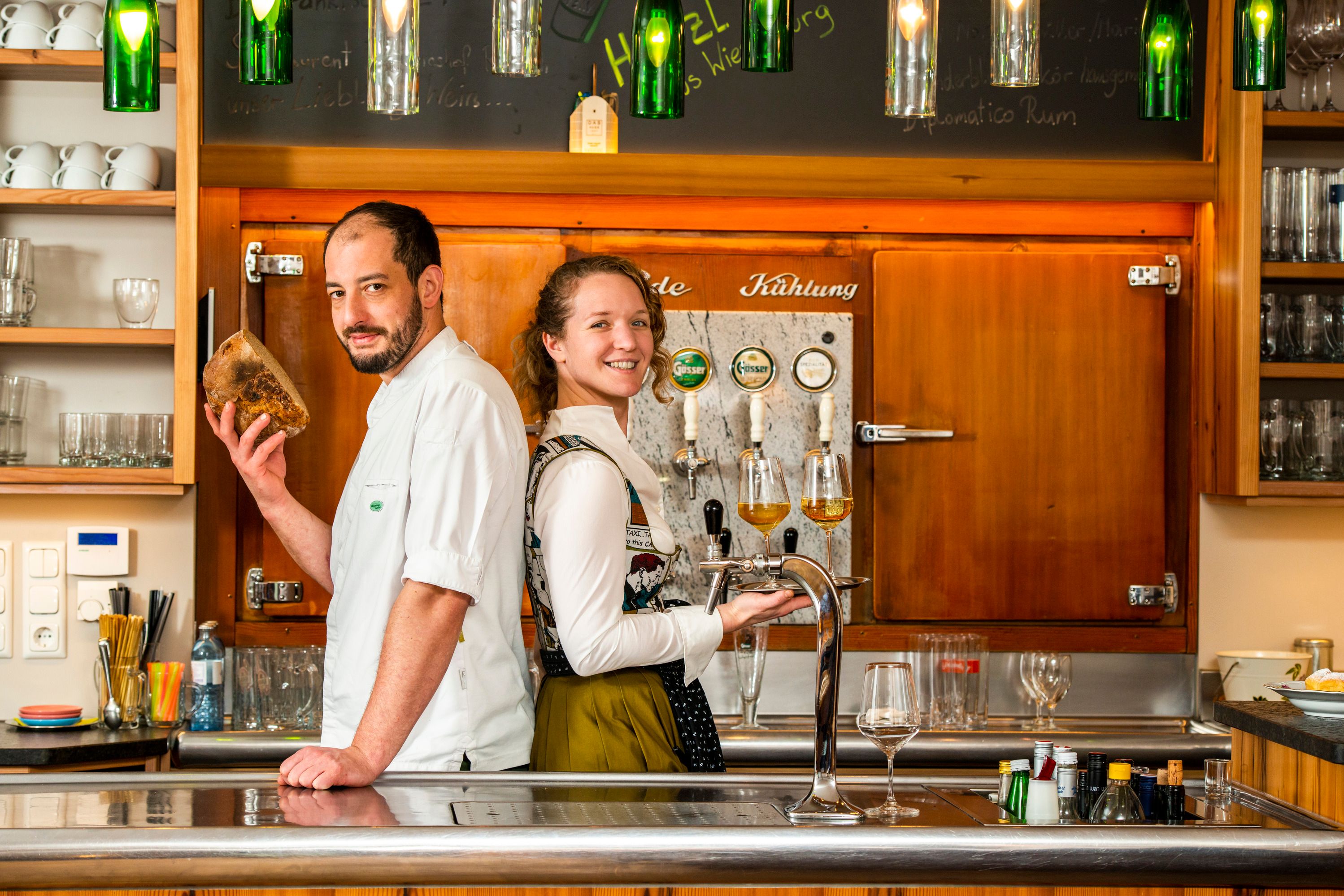 Zwei Personen stehen in einer Bar, eine hält Brot, die andere ein Tablett mit Getränken.