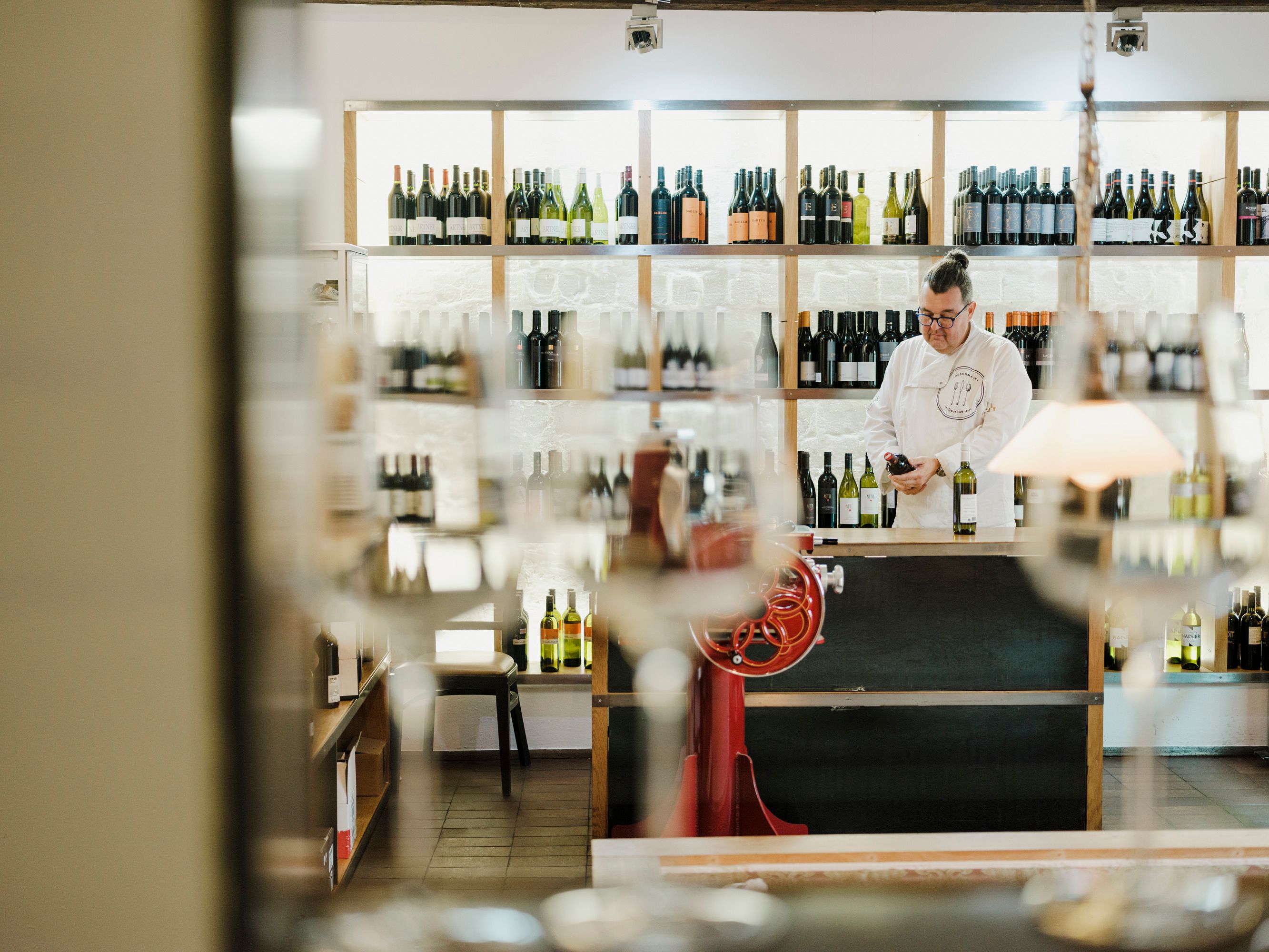 Mann in Vinothek mit Regalen voller Weinflaschen, unscharfe Gläser im Vordergrund.