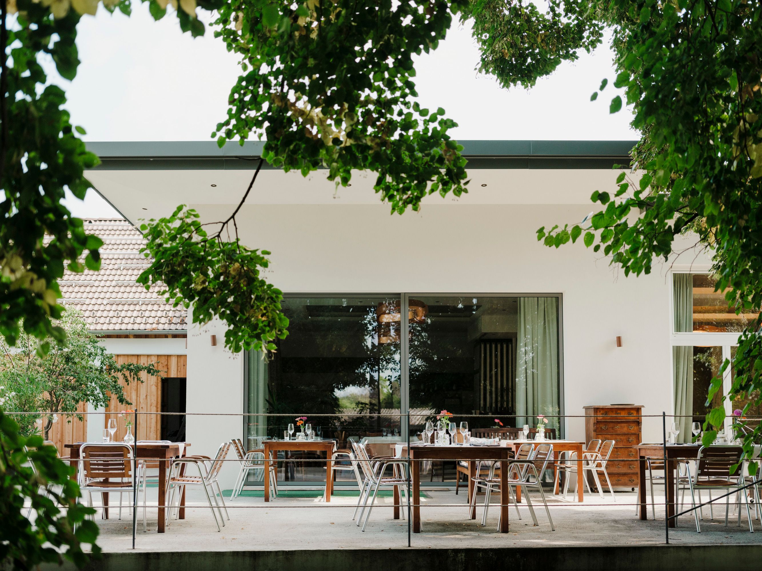 Terrasse mit Tischen und Stühlen, umgeben von grünen Bäumen.