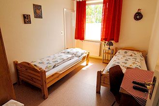 Ein einfach eingerichtetes Zimmer mit zwei Einzelbetten, einem Fenster mit roten Vorhängen und einem kleinen Tisch mit Stuhl.