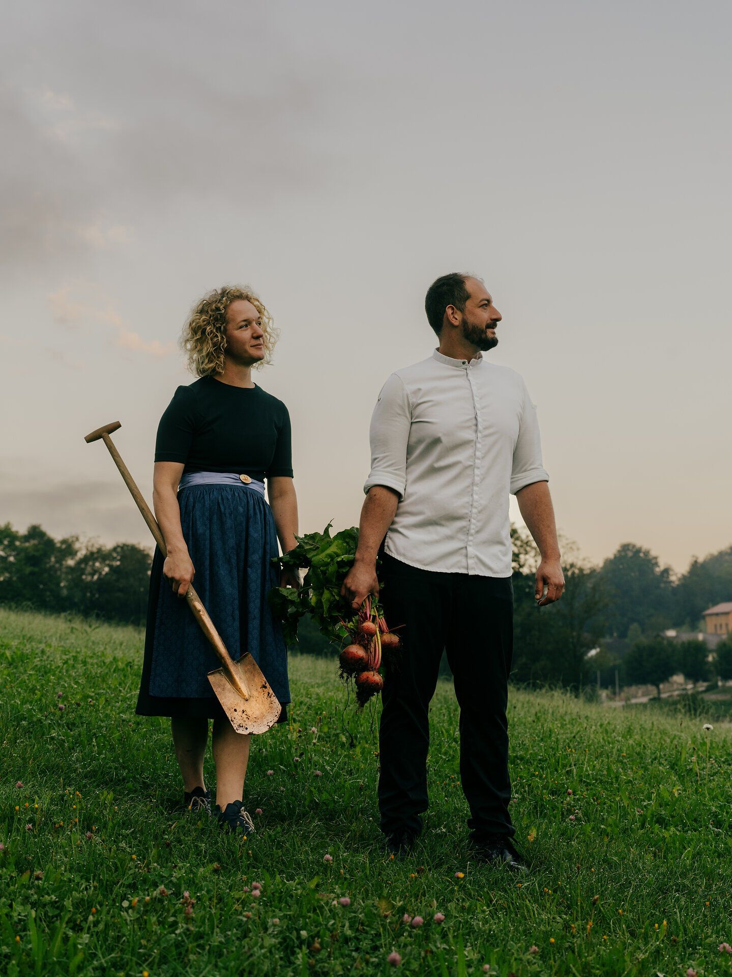 In der sanften Abenddämmerung stehen zwei Personen auf einer grünen Wiese, umgeben von der idyllischen Landschaft des Mostviertels. Der Mann hält frisch geerntetes Gemüse in der Hand, während die Frau mit einer Schaufel bereit ist, die Erde zu bearbeiten. Diese Szene strahlt eine harmonische Verbindung zur Natur und zur regionalen Landwirtschaft aus.