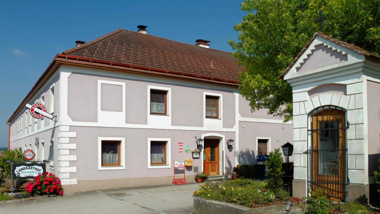 Ein traditioneller Landgasthof mit grauer Fassade und roten Dachziegeln, umgeben von Blumen und Bäumen.