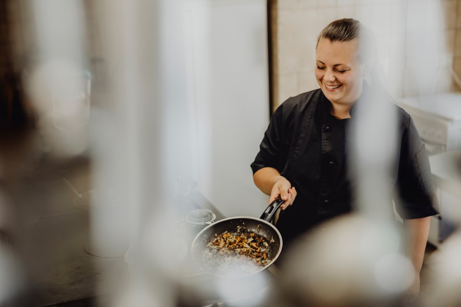 Eine K&ouml;chin in schwarzer Uniform h&auml;lt eine Pfanne mit gebratenem Gem&uuml;se und l&auml;chelt.