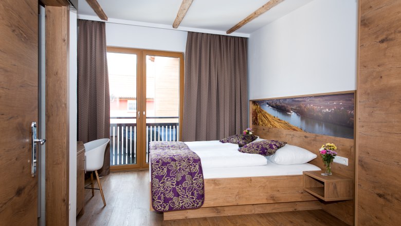 Ein gem&uuml;tliches Hotelzimmer mit Holzm&ouml;beln, einem Doppelbett, Balkon und dekorativen Kissen.