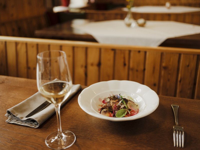 Ein Glas Wei&szlig;wein und ein Teller mit Gourmetgericht auf einem Holztisch in einem rustikalen Restaurant.