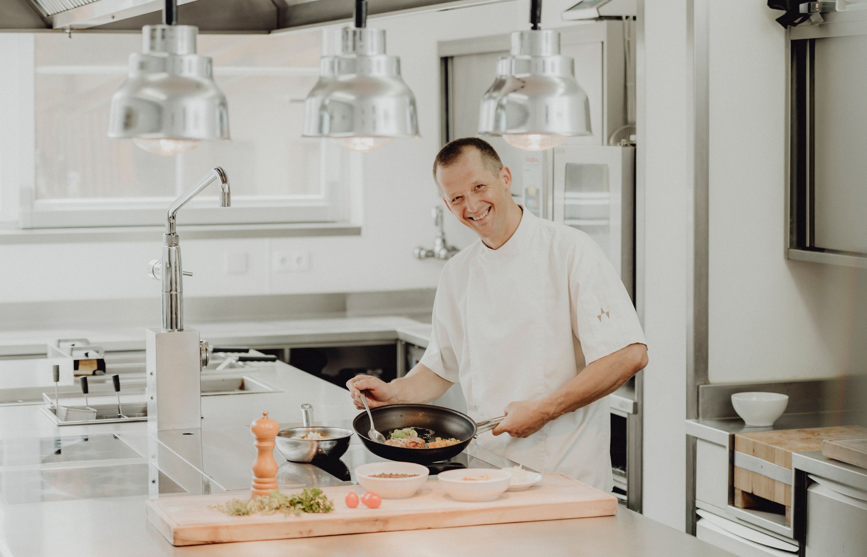 Ein Koch in einer modernen Küche, der lächelnd eine Pfanne hält.