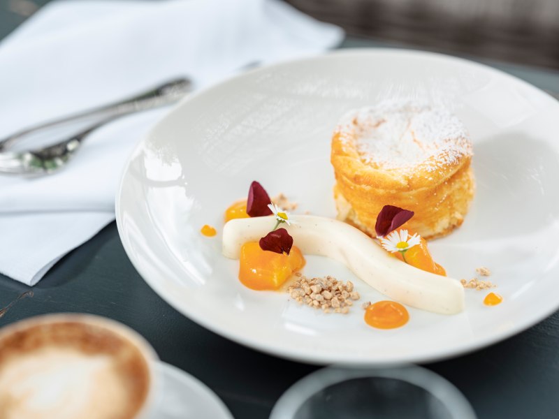 Ein Dessertteller mit Topfenstrudel-Souffl&eacute;, Vanillecreme, Fruchtso&szlig;e und essbaren Bl&uuml;ten, daneben ein Cappuccino.