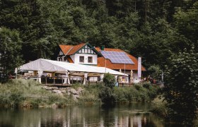 Gasthaus mit Solarpanelen und Zelt am Waldrand neben einem Teich.