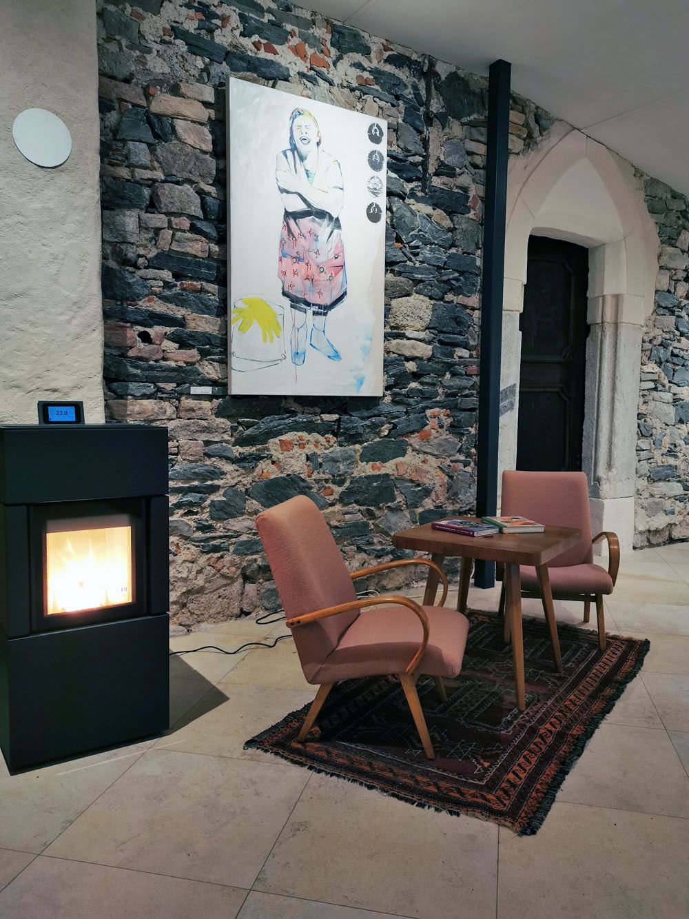 Gemütlicher Raum mit Kamin, zwei Sesseln, Tisch und Kunstwerk an einer Steinwand.