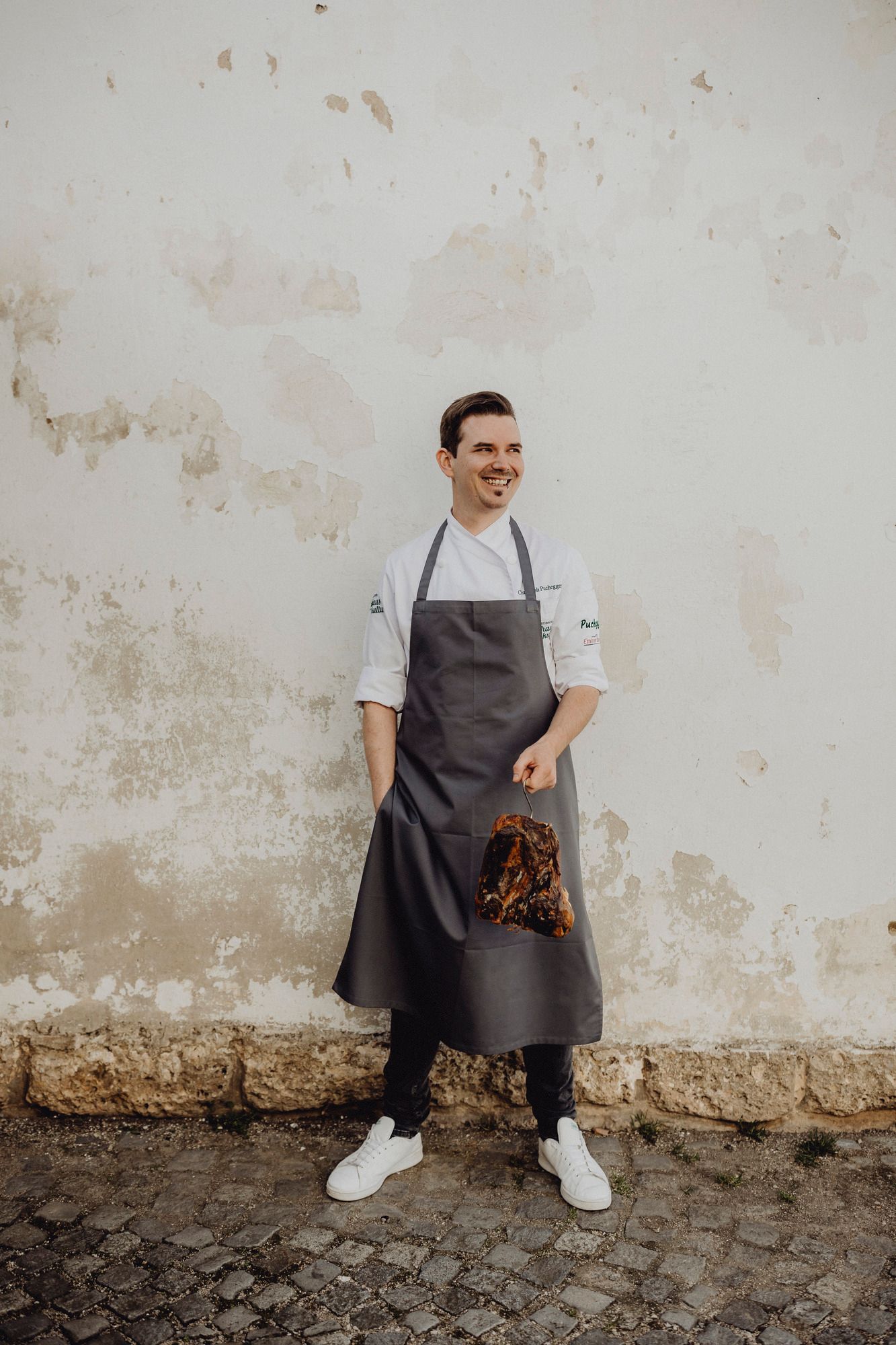 Ein Koch in weißem Hemd und grauer Schürze hält ein großes Stück Fleisch vor einer alten Wand.