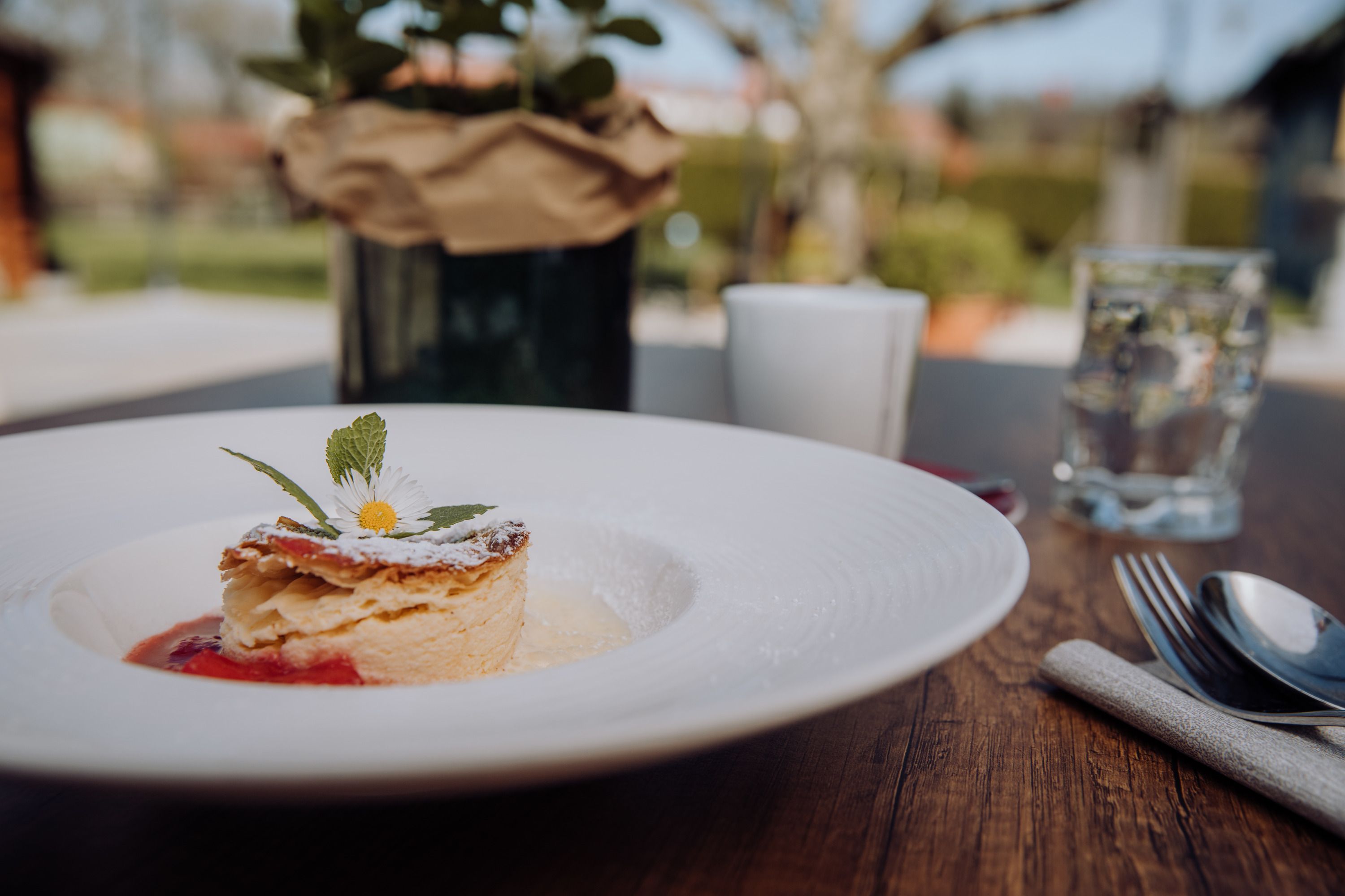 Ein Teller mit Milchrahmstrudel, garniert mit einem Gänseblümchen, auf einem Holztisch im Freien.