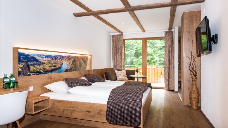 Modernes Hotelzimmer mit Holzm&ouml;beln, gro&szlig;em Bett und Balkon mit Blick ins Gr&uuml;ne.
