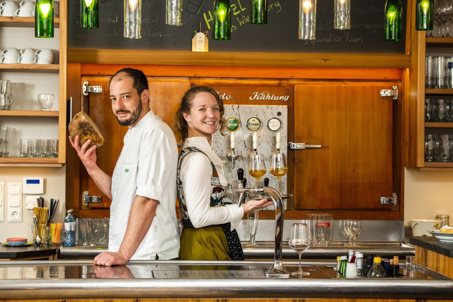 Zwei Personen stehen in einer Bar, eine hält Brot, die andere ein Tablett mit Getränken.