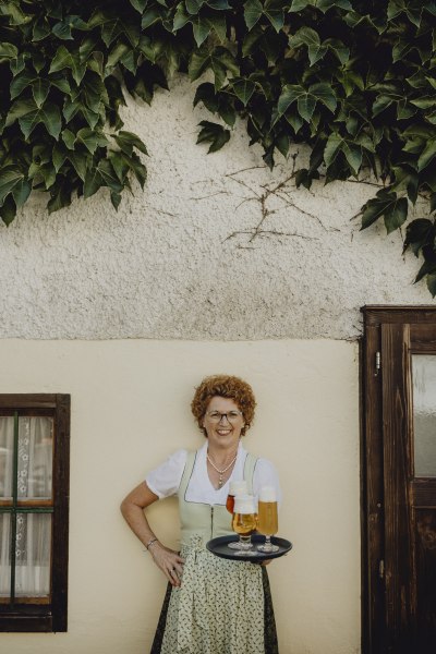 Eine Frau in traditioneller Kleidung h&auml;lt ein Tablett mit Getr&auml;nken vor einer mit Efeu bewachsenen Wand.
