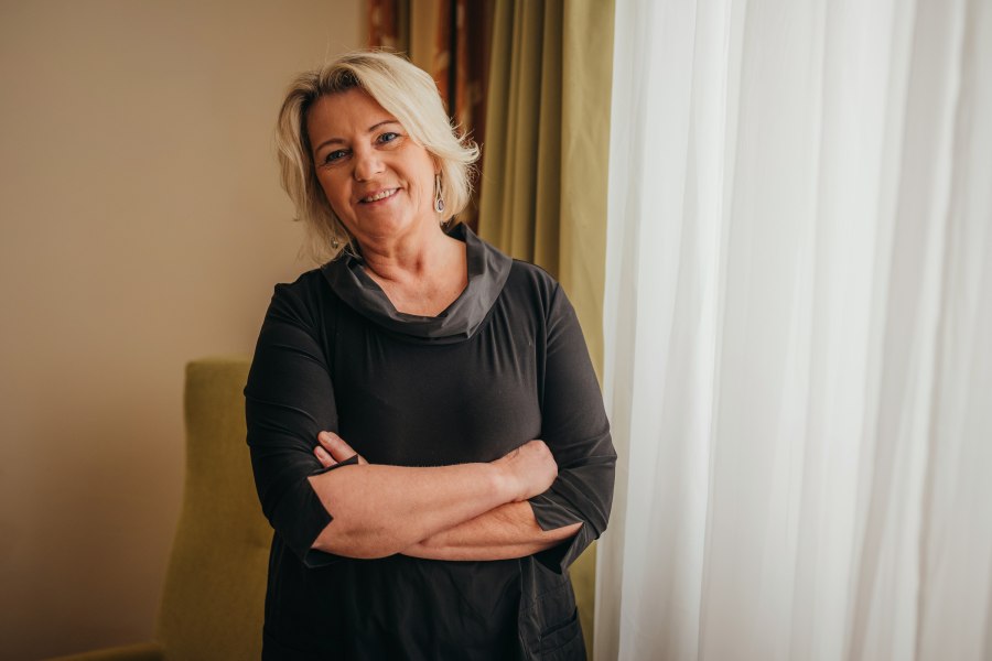 Eine Frau mit blonden Haaren steht l&auml;chelnd vor einem Fenster mit Vorh&auml;ngen.