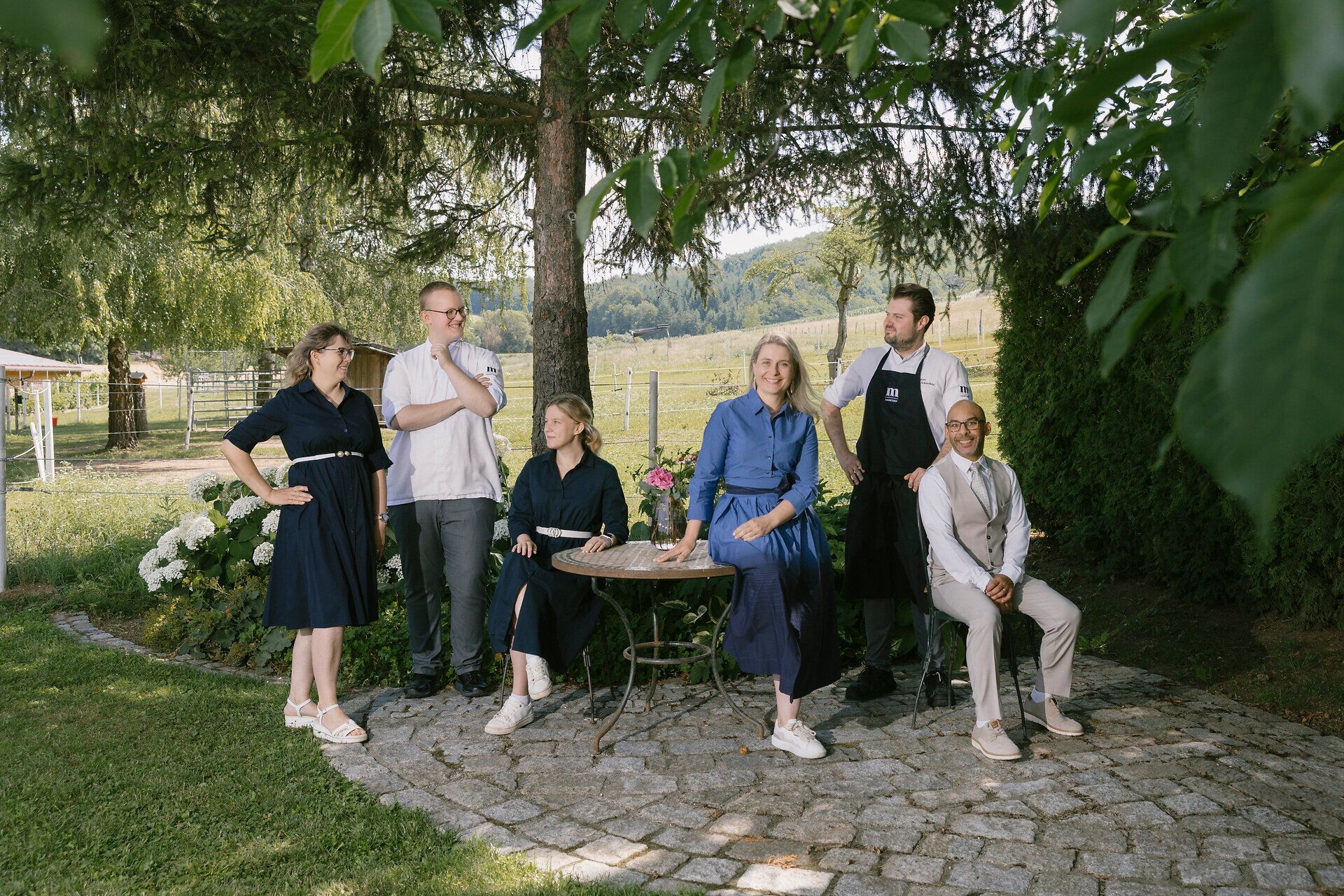 Sechs Personen stehen und sitzen gemeinsam im Garten unter Bäumen rund um einen kleinen runden Tisch. Im Hintergrund sind Wiese, Zaun, Bäume und Hügel zu sehen.