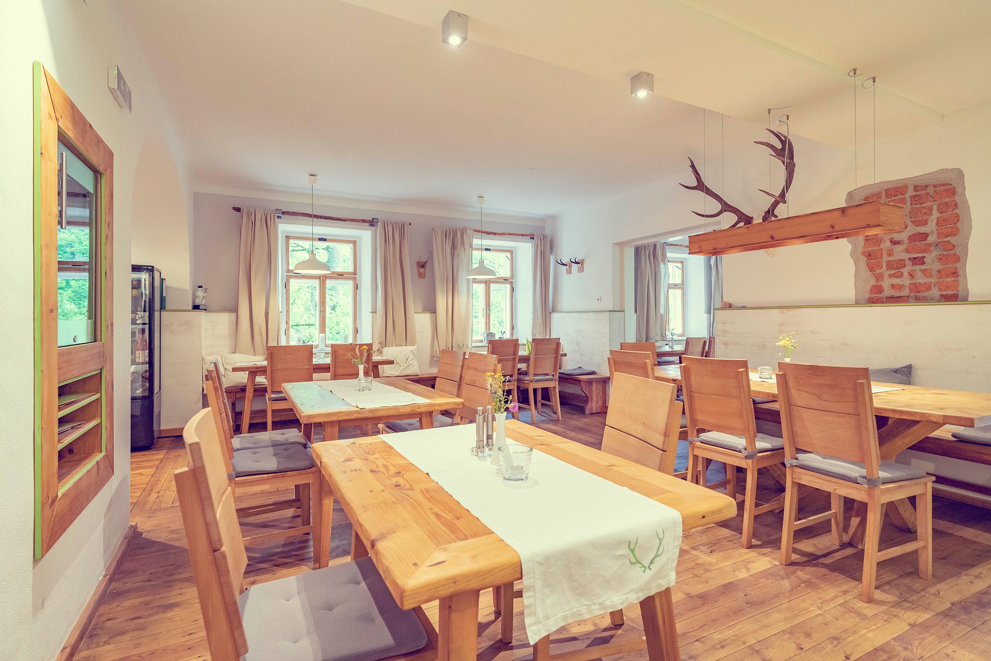 Ein rustikal-moderner Raum mit Holzmöbeln, weißen Wänden und großen Fenstern. Ein Geweih hängt als Dekoration von der Decke.