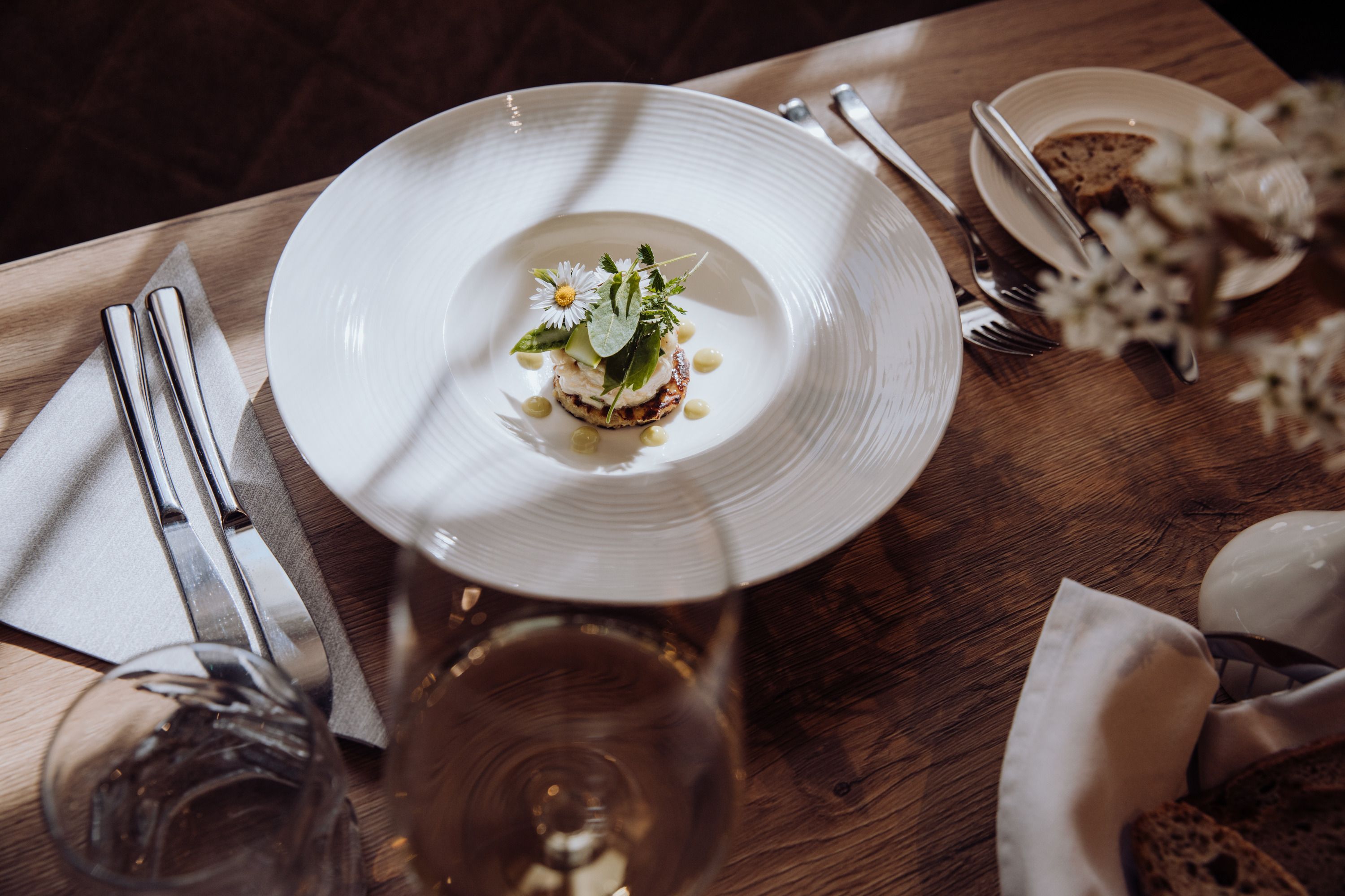 Ein Teller mit Tartar von Räucherforelle auf einem Erdäpfelpuffer, garniert mit Kräutern und Blüten, auf einem Holztisch mit Besteck und einem Glas Wein.