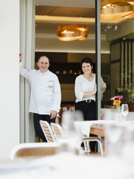 Zwei Personen stehen in einem Restaurant am Eingang, eine in Kochkleidung, die andere in wei&szlig;er Bluse.