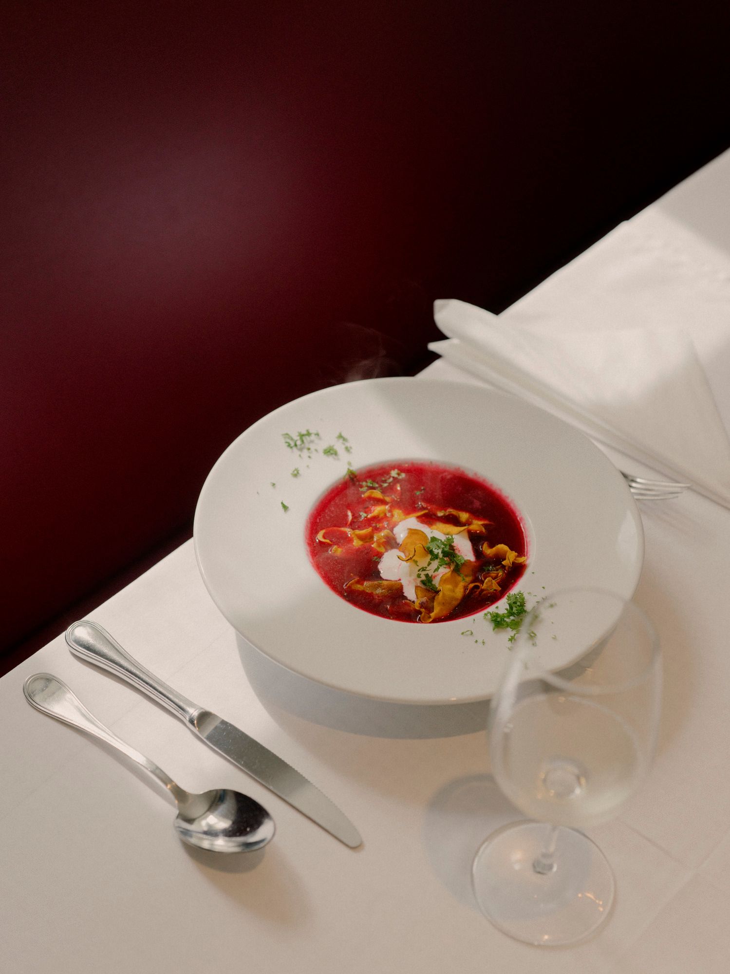 Ein Teller mit roter Rübensuppe, garniert mit Sahne und Kräutern, auf einem gedeckten Tisch mit Besteck und einem Weinglas.