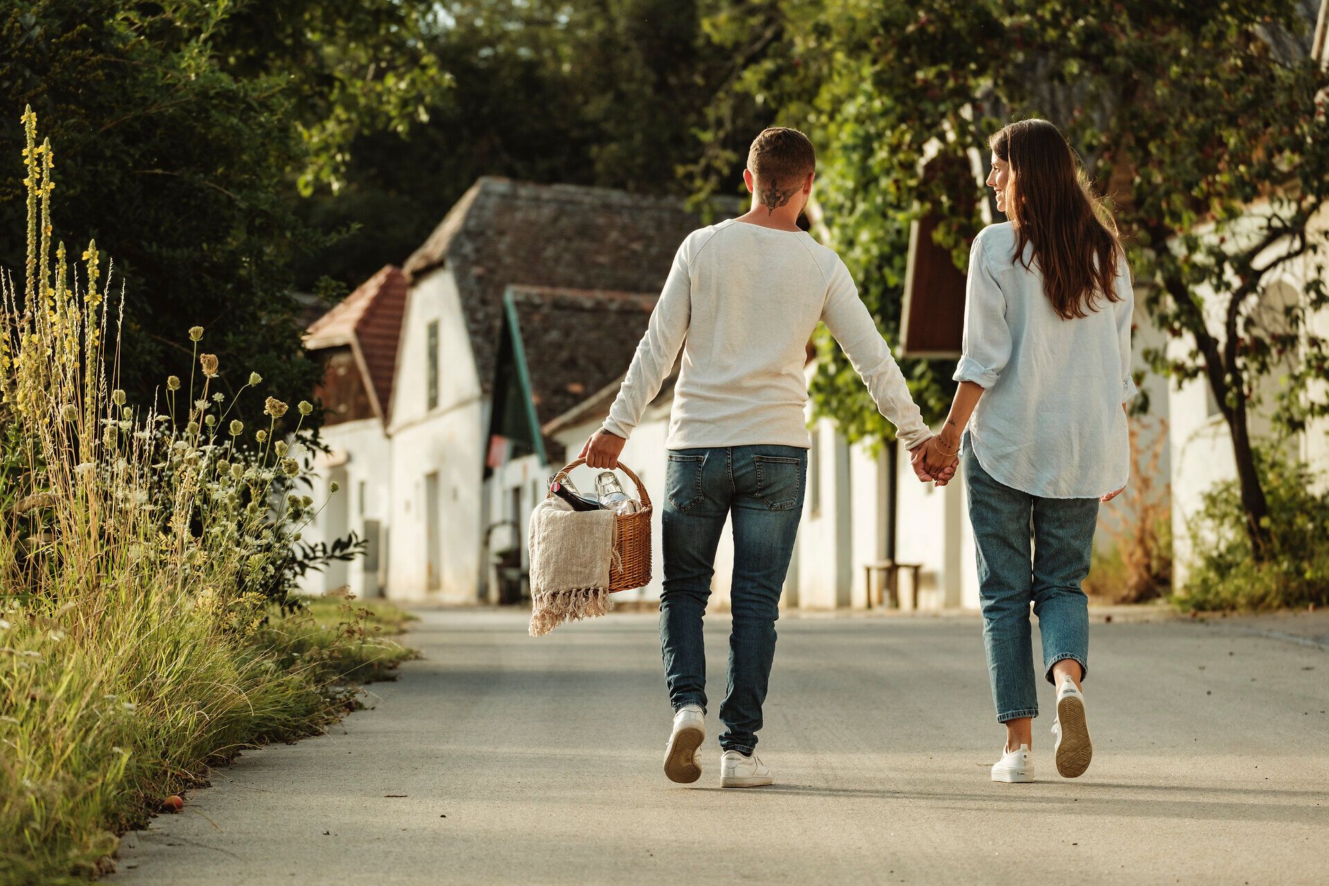 Ein Paar genießt einen romantischen Spaziergang durch die malerische Kellergasse, während sie Hand in Hand einen Picknickkorb tragen. Umgeben von üppigem Grün und charmanten Weinhäusern, strahlt die Atmosphäre pure Lebensfreude aus. Der Duft von frischem Wein und köstlichen Snacks erfüllt die Luft und lädt zum Verweilen ein.