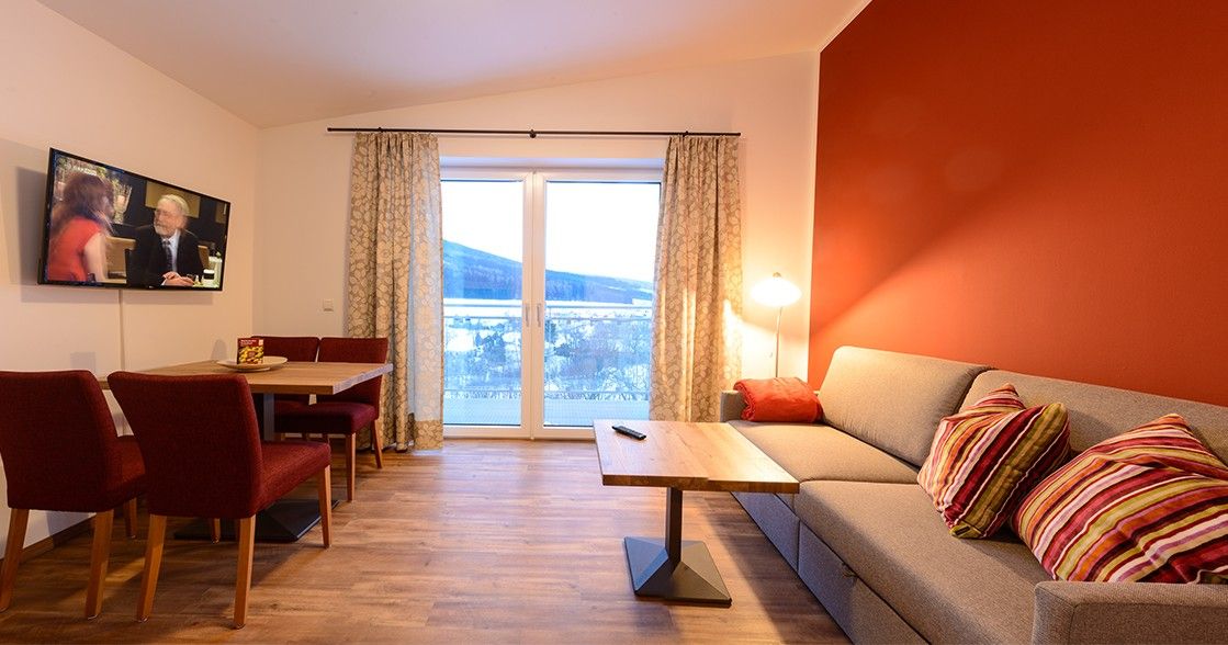 Gemütliches Wohnzimmer mit roter Akzentwand, grauem Sofa, Holztisch, Fernseher und großem Fenster mit Vorhängen.