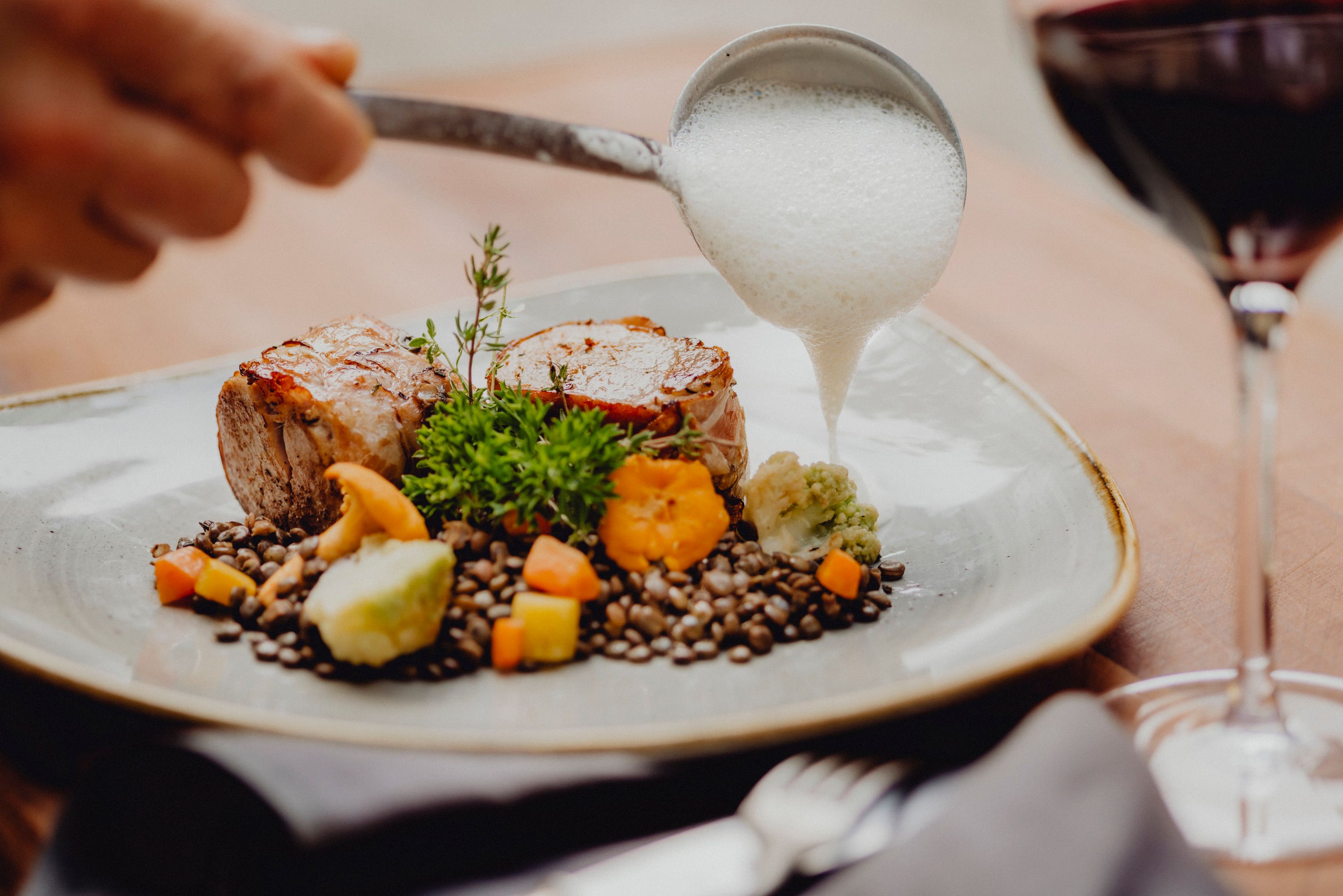 Schweinemedaillons mit Speckmantel, Linsen und Veltliner-Schaum auf einem Teller, daneben ein Glas Rotwein.