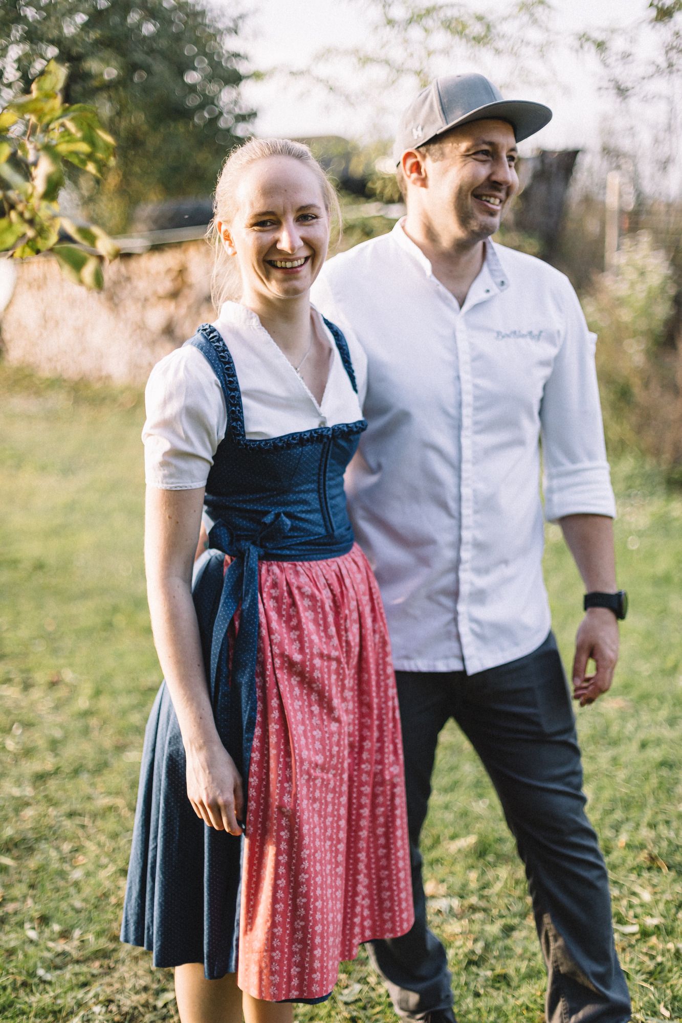Ein Mann und eine Frau stehen lächelnd im Freien. Die Frau trägt ein traditionelles Dirndl, der Mann ein weißes Hemd und eine Kappe.