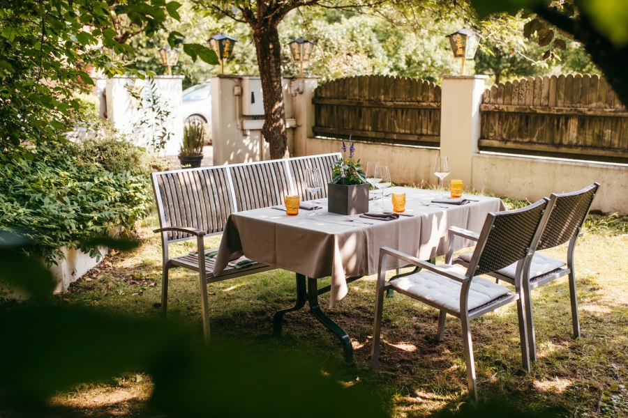 Gedeckter Tisch im Garten mit St&uuml;hlen und B&auml;nken unter B&auml;umen.