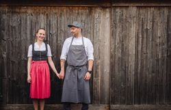 In der warmen Umarmung des Mostviertels strahlen die Wirtsleut in traditioneller Tracht, während sie Hand in Hand stehen und die herzliche Gastfreundschaft verkörpern. Die rustikale Holzfassade des Wirtshauses im Hintergrund erzählt von unzähligen Geschichten und kulinarischen Genüssen, die hier erlebt werden können.