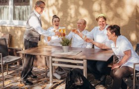 Ein Team von fünf Personen stößt draußen an einem Tisch an, mit einem schwarzen Hund im Vordergrund.