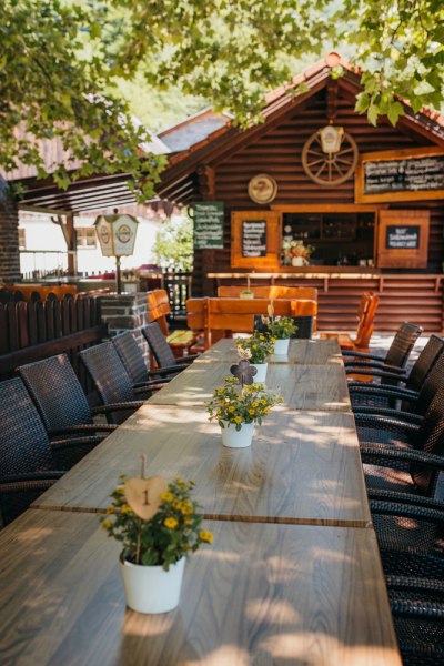 Ein langer Holztisch im Freien mit Blumen in T&ouml;pfen, umgeben von St&uuml;hlen. Im Hintergrund eine Holzh&uuml;tte mit Schildern.
