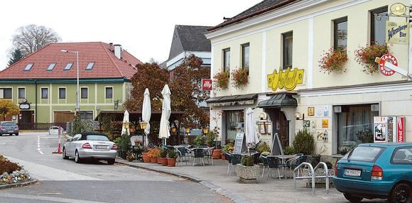 Außenansicht von einem Gasthaus an einem Belebten Platz mit Bäumen, Sonnenschirmen, Sesseln uvm.
