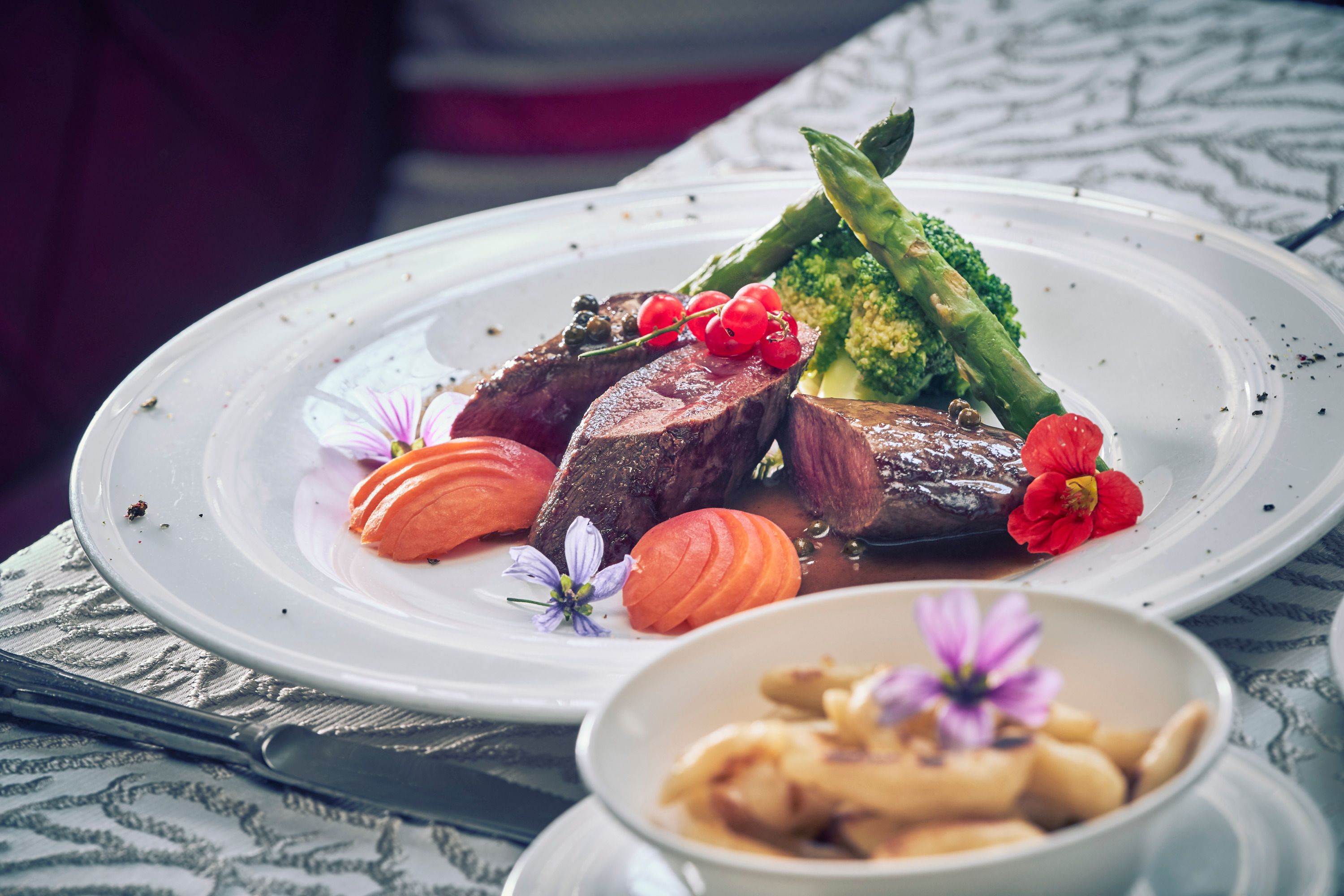 Ein Teller mit Rehrücken, Pfeffer-Marillen, Brokkoli und Spargel, dekoriert mit Blumen. Im Vordergrund eine Schüssel mit Schupfnudeln.