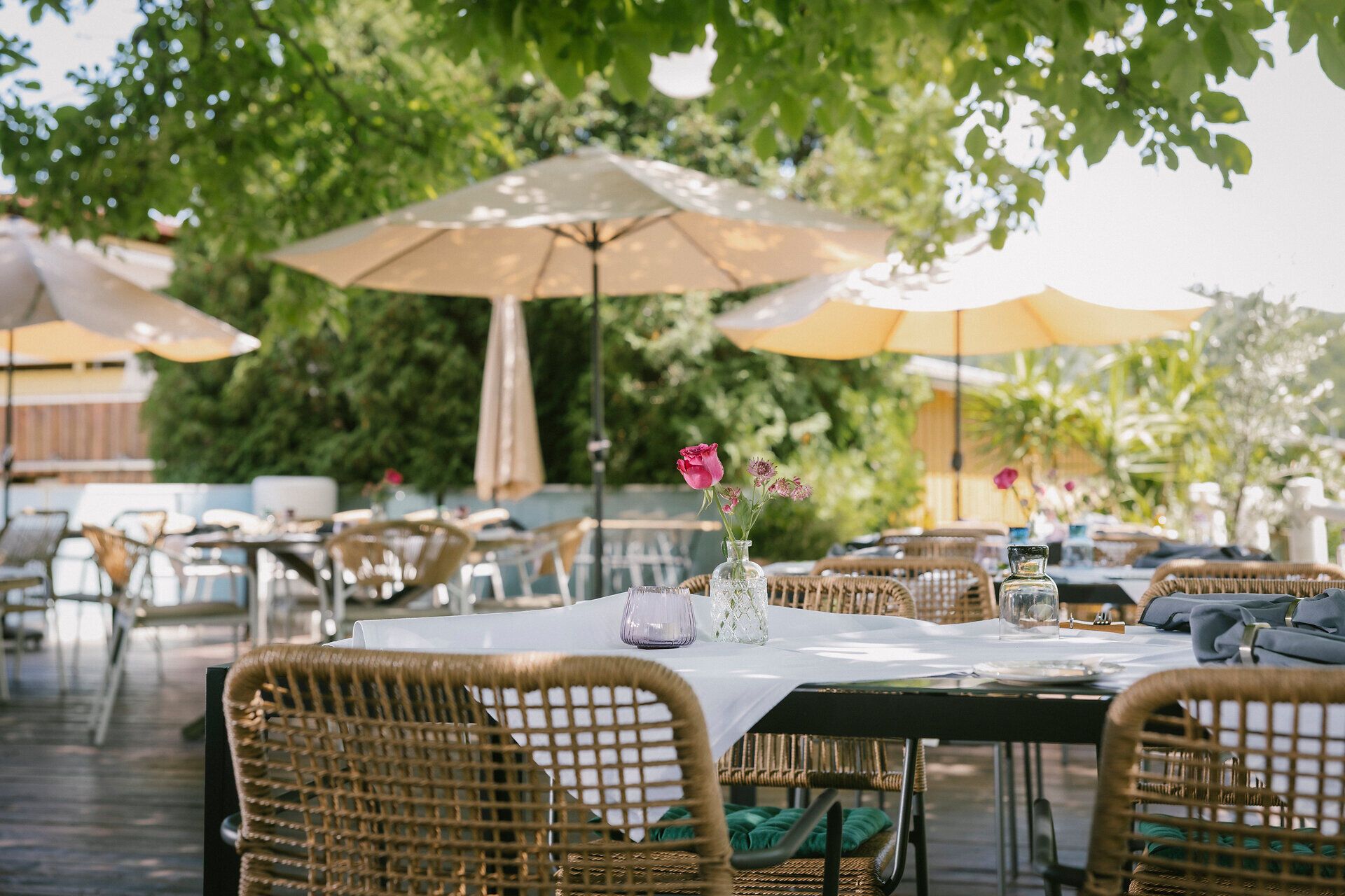 Gedeckte Tische mit Korbstühlen stehen auf einer Terrasse unter großen Sonnenschirmen und Bäumen. Auf den Tischen stehen Blumenvasen, Gläser und Geschirr.