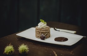 Ein Dessert aus Kastanienmousse auf einem wei&szlig;en Teller, garniert mit Sahne und Minzblatt, daneben eine Schokoladensauce und eine Kirsche.