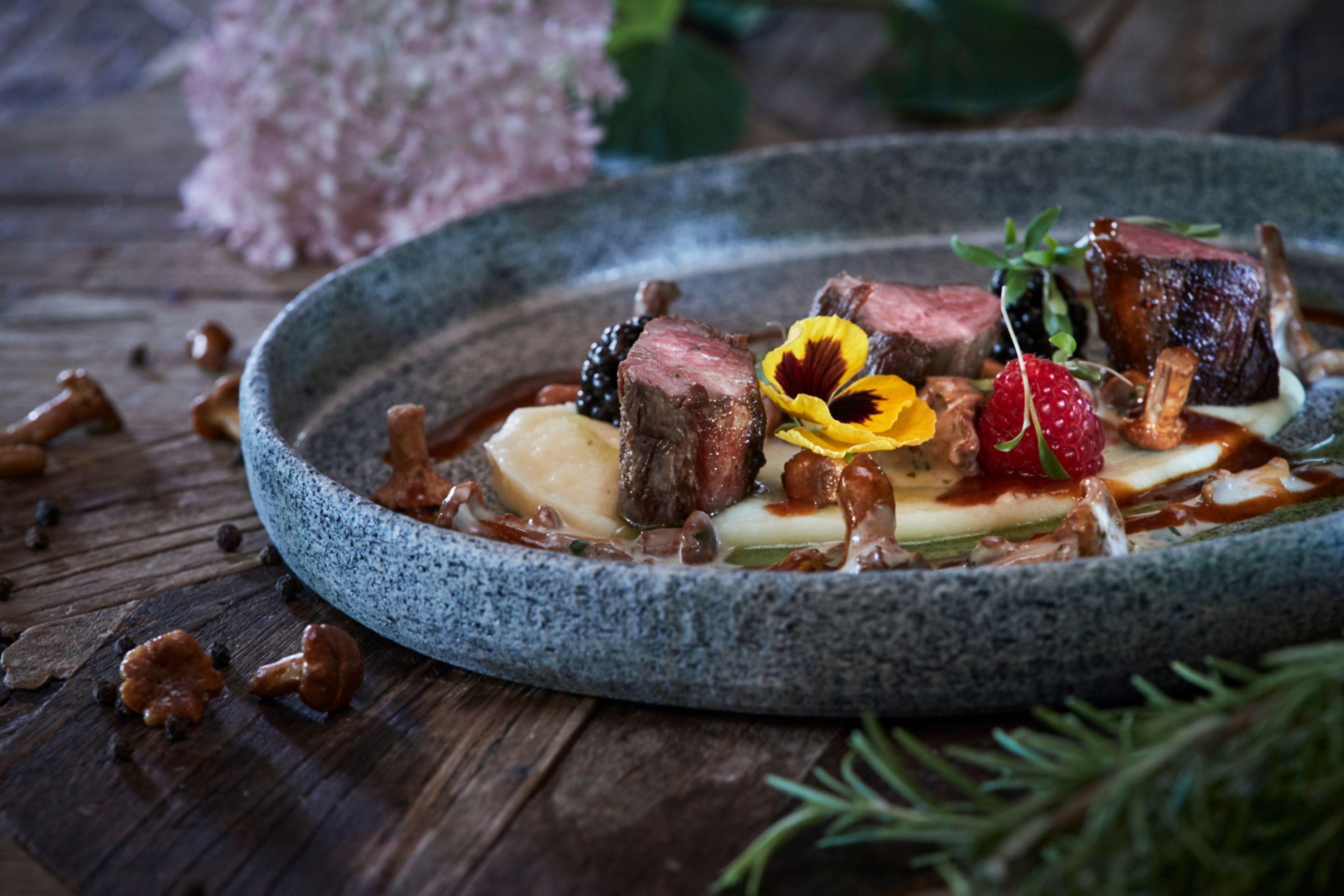 Ein Teller mit Rehfilet, Eierschwammerln, Beeren und essbaren Blüten auf einem Holztisch.
