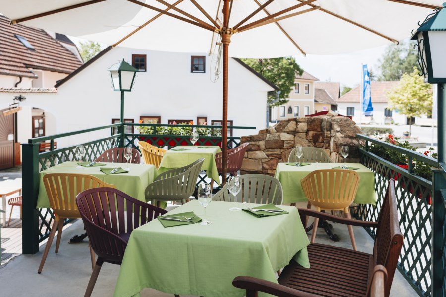 Ein Gastgarten mit gr&uuml;nen Tischdecken und bunten St&uuml;hlen unter einem Sonnenschirm, umgeben von Geb&auml;uden und Pflanzen.