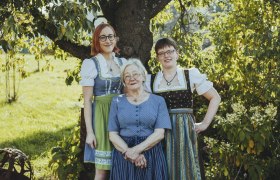 Drei Frauen in traditioneller Tracht stehen l&auml;chelnd unter einem Baum im Freien.