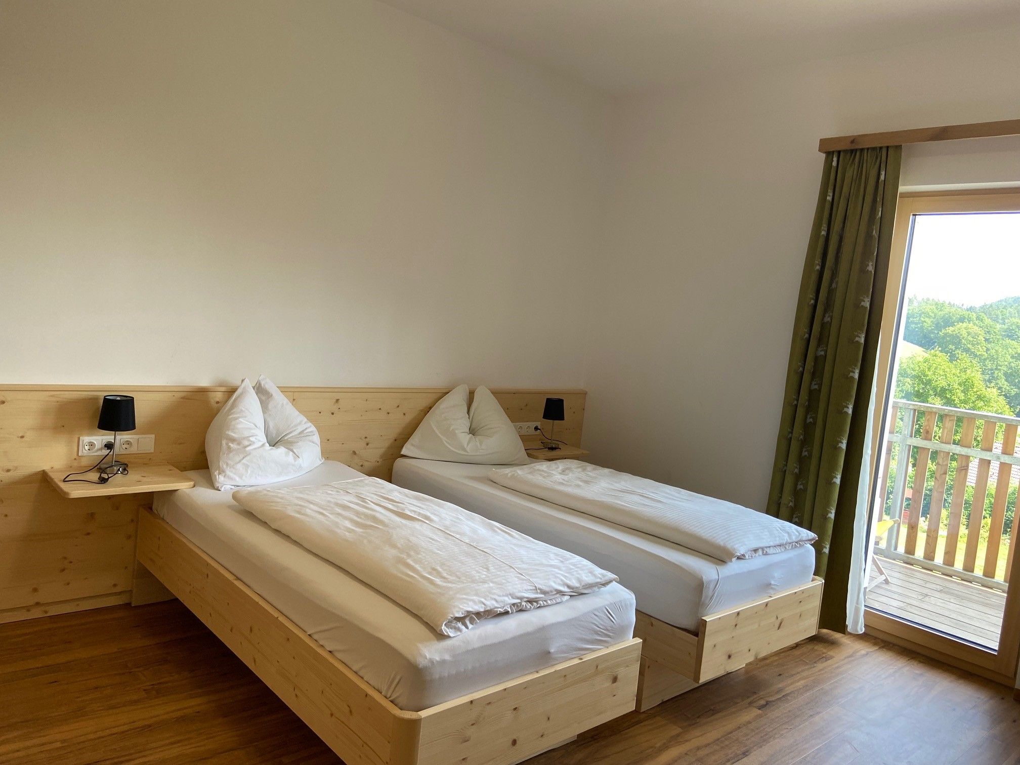 Zweibettzimmer mit Holzboden, zwei Einzelbetten, Nachttischlampen und einem Balkon mit Ausblick ins Grüne.