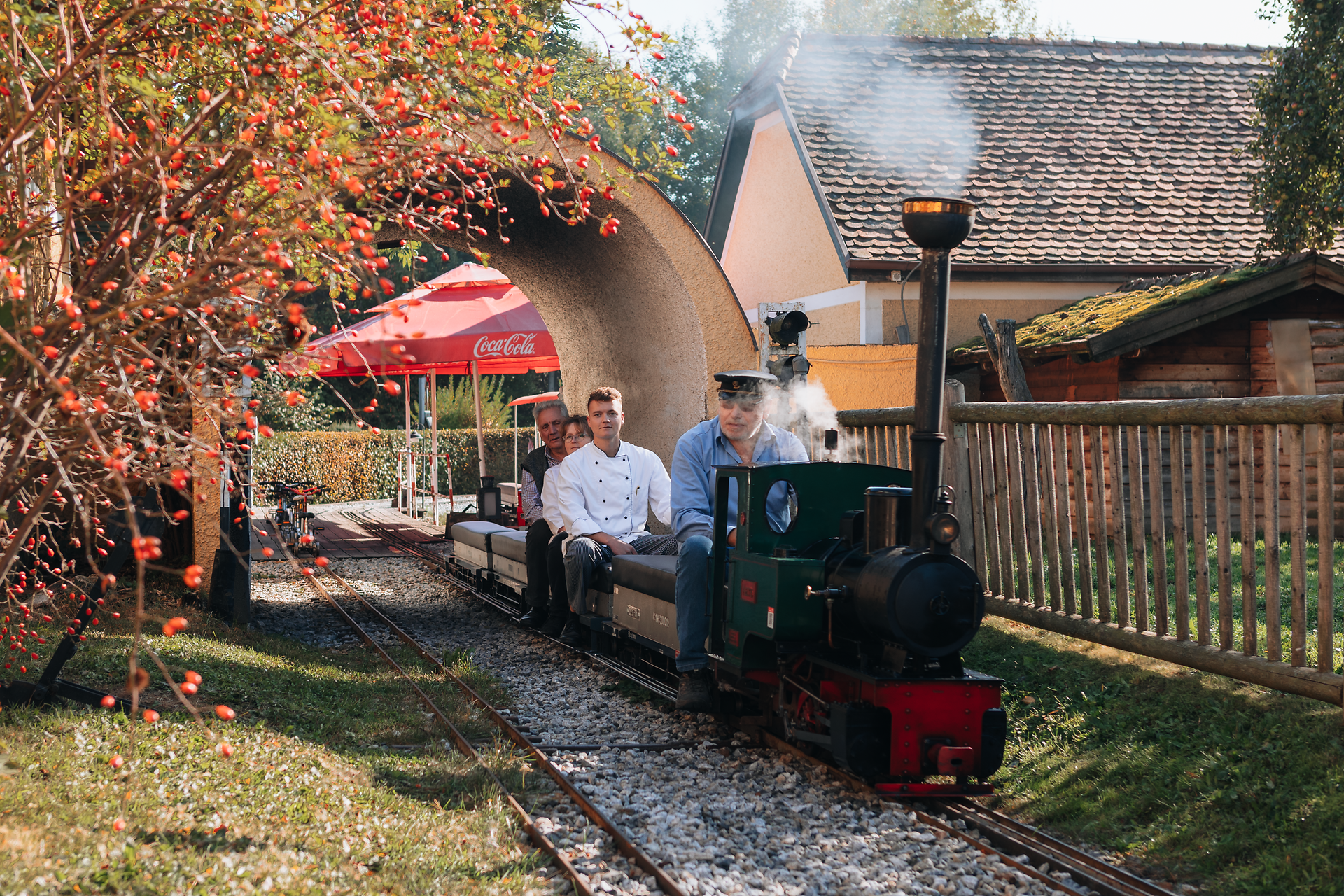 Eine kleine Dampflok schnauft fröhlich durch die malerische Landschaft, während die Passagiere in festlicher Stimmung die Fahrt genießen. Umgeben von bunten Bäumen und dem sanften Plätschern eines Baches, vermittelt dieser Ort ein Gefühl von unbeschwerter Freude und kindlicher Neugier.