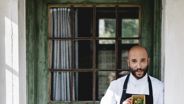 Ein Mann in Kochkleidung hält eine Schachtel mit Kräutern vor einer grünen Holztür.