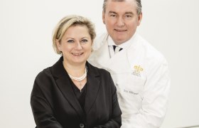 Ein Mann und eine Frau posieren lächelnd vor einem weißen Hintergrund.