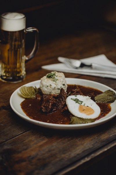 Ein Teller mit Gulasch, Spiegelei, Kn&ouml;del und Gurken auf einem Holztisch, daneben ein Bierkrug.