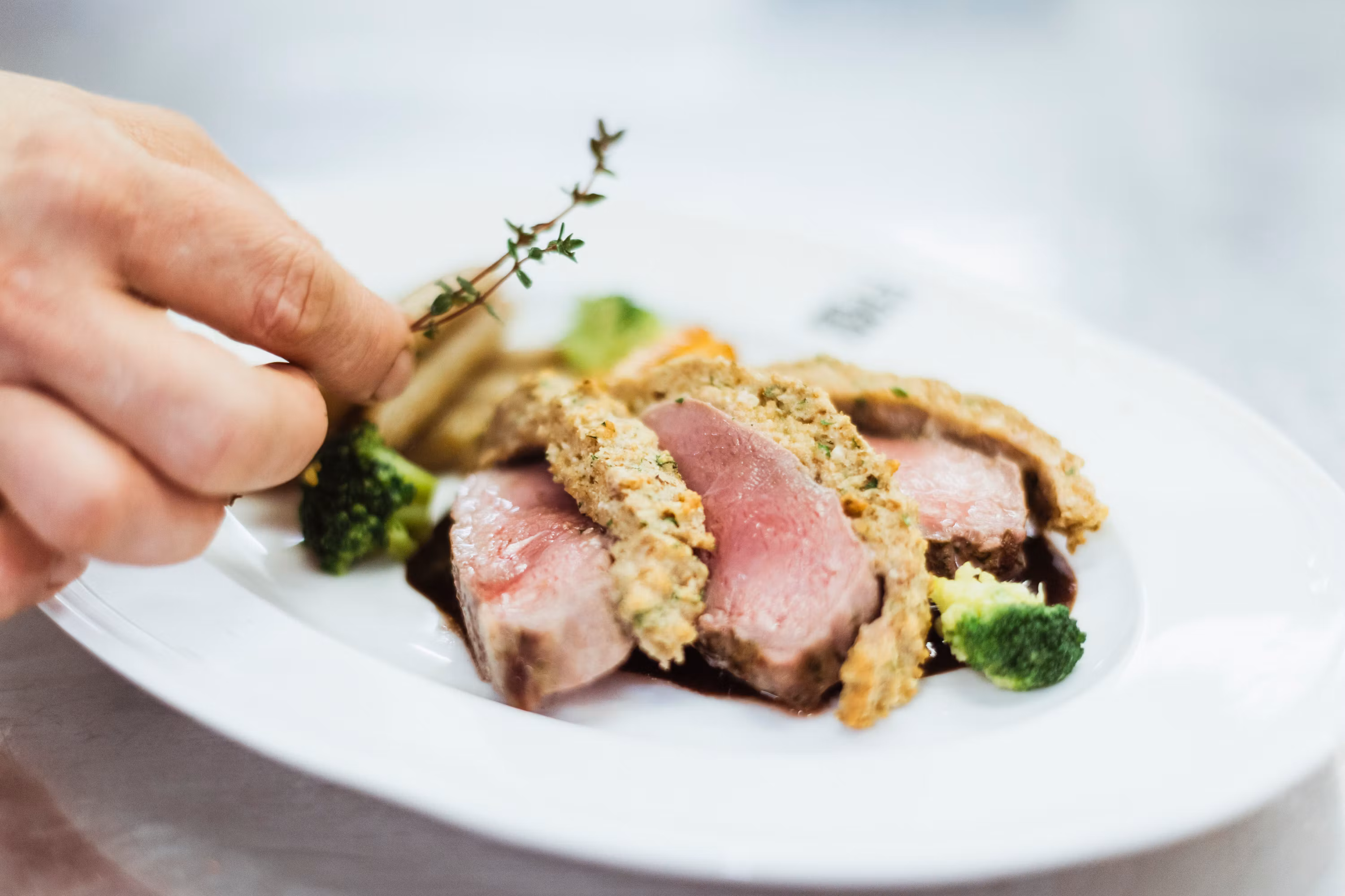 Nahaufnahme eines rosa gebratenen Fleischgerichts mit Kräuterkruste, Brokkoli und einer Hand, die Thymian hinzufügt.