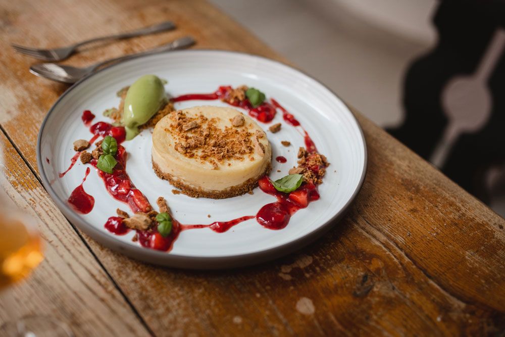 Ein Teller mit einem Cheesecake, dekoriert mit roten Beeren und grünem Eis, auf einem Holztisch.