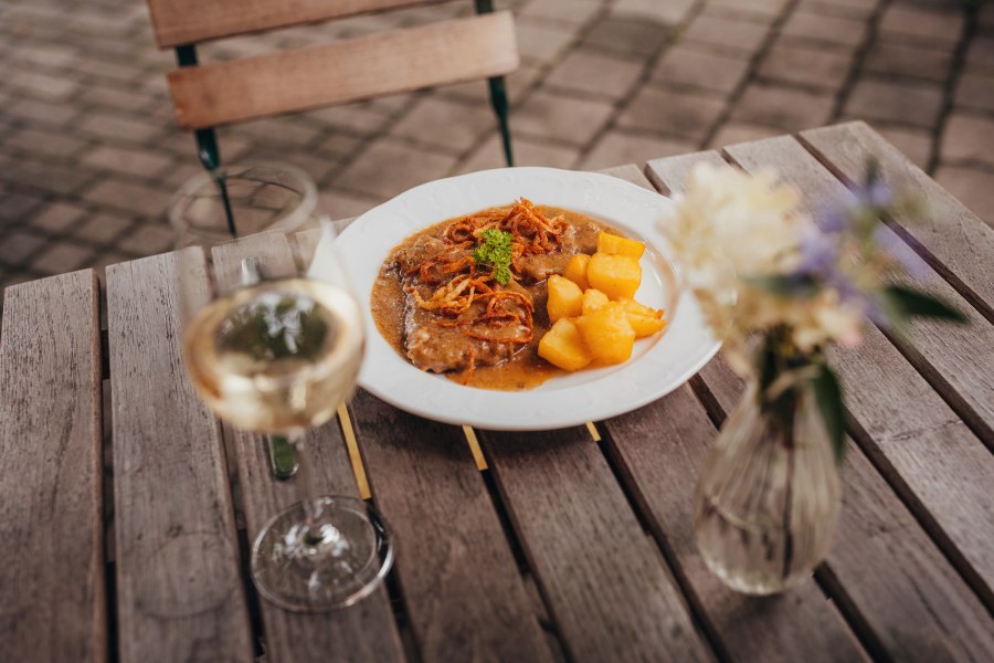 Ein Teller mit Zwiebelrostbraten und Braterd&auml;pfeln auf einem Holztisch, daneben ein Glas Wei&szlig;wein und eine Blumenvase.