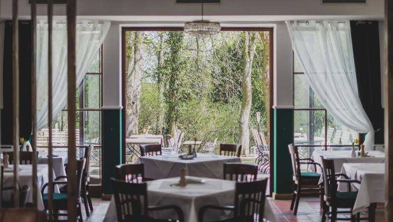 Ein Restaurant mit gedeckten Tischen und Blick auf einen gr&uuml;nen Garten durch gro&szlig;e Fenster.