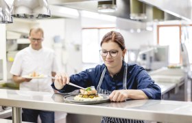 Köchin in einer Restaurantküche richtet ein Gericht an, während ein Kollege im Hintergrund ein weiteres Gericht hält.