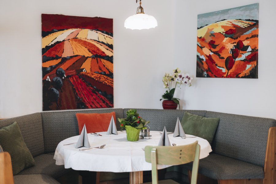 Gem&uuml;tliche Sitzecke mit Tisch, dekoriert mit grauen Servietten und einer Pflanze, umgeben von bunten Landschaftsgem&auml;lden an der Wand.